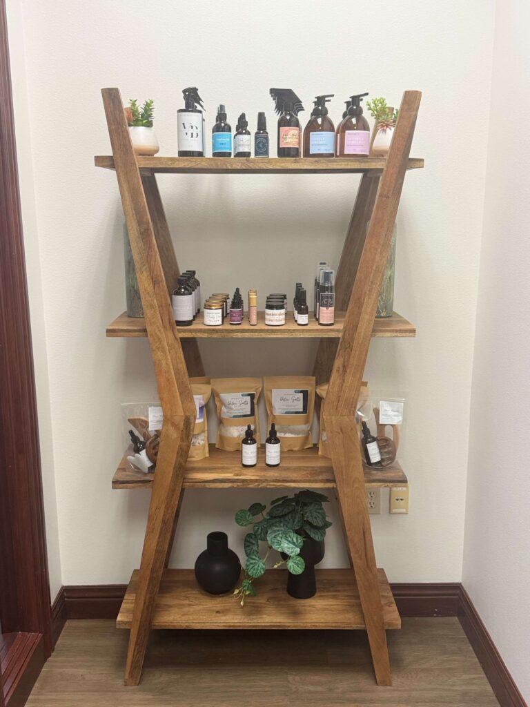 A wooden shelf displays various skincare and wellness products, including oils, sprays, and herbal packages, alongside decorative plants.